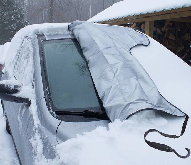 The cover, partially removed from a windshield of a car covered in snow, showing how the windshield is totally snow-free under the cover