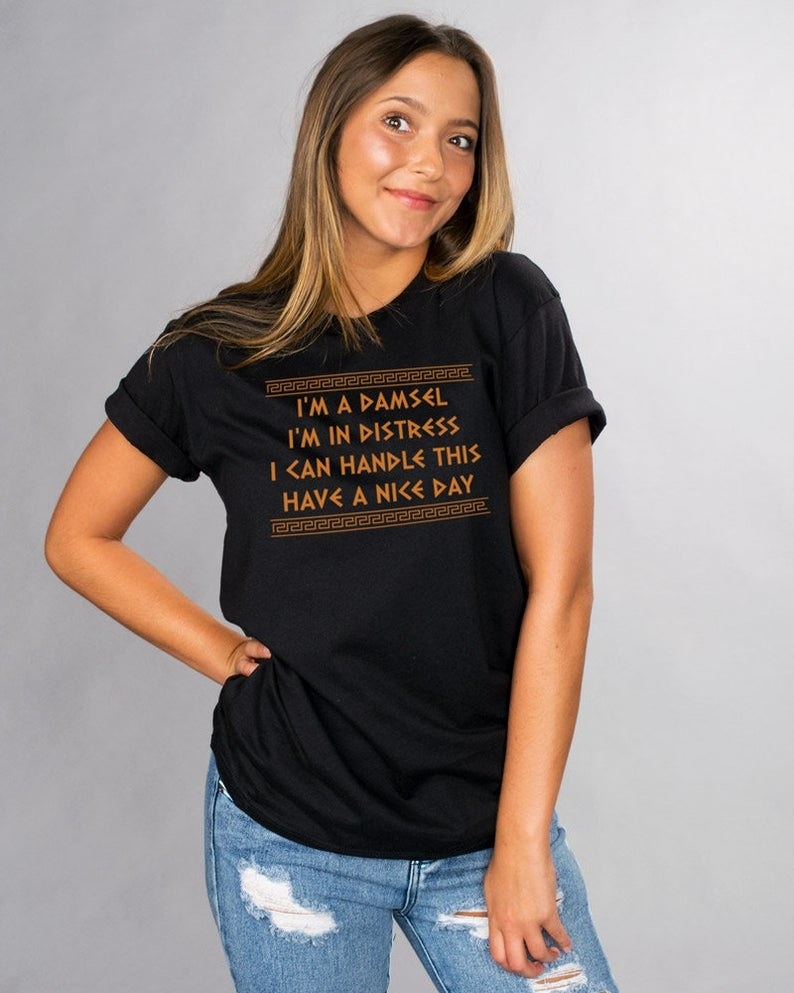 Model wearing the t-shirt in black that says &quot;I&#x27;m a damsel I&#x27;m in distress I can handle this Have a nice day&quot; in gold