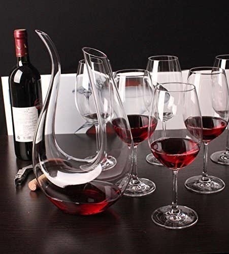 A u-shaped glass wine decanter with red wine in it and wine glasses and a bottle and opener around it
