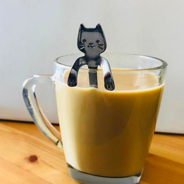 The spoon, which has a cat face and bent arms at the top of the handle, resting on a mug of coffee