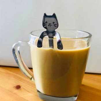 The spoon, which has a cat face and bent arms at the top of the handle, resting on a mug of coffee