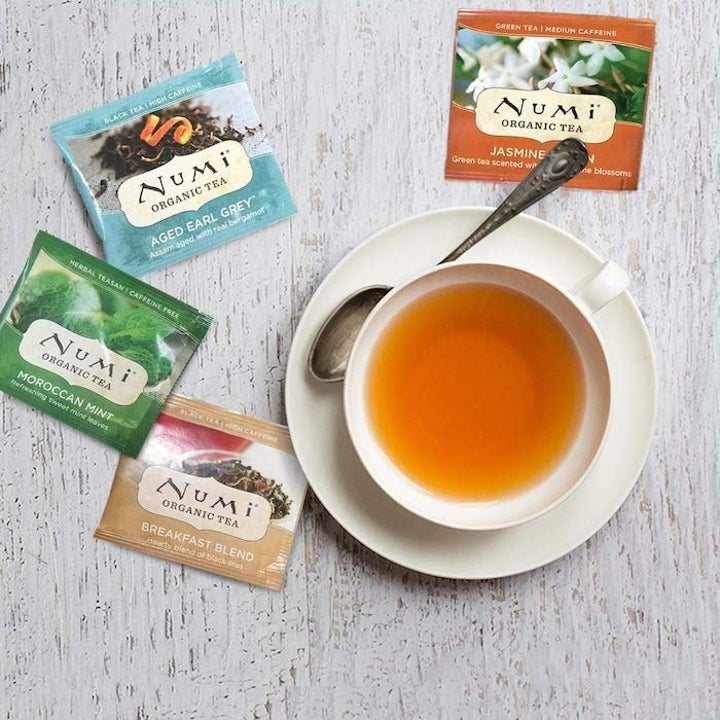 This is a photo of a white saucer holding a teacup full of orange tea and a spoon. There are four tea packets sitting next to the saucer in different colors.