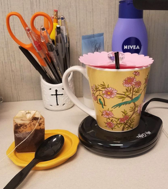 A mug sitting on top of the Mr. Coffee mug warmer, which looks like a thick coaster.  There's also a spoon and a tea bag sitting on an actual coaster right beside it. This warmer is sitting on a desk.