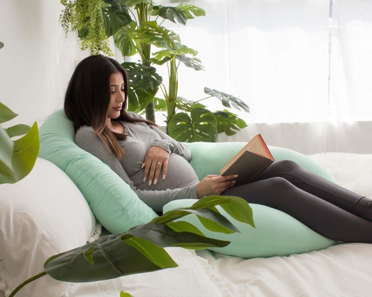 Pregnant person reading book in bed propping back and legs up with large c-shaped pillow 