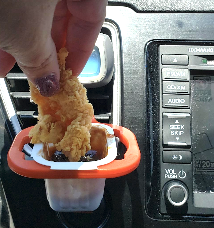 the sauce dipper attached to the vent in the car and a reviewer&#x27;s hand dumping a crispy chicken tender into a pack of sauce sitting in it