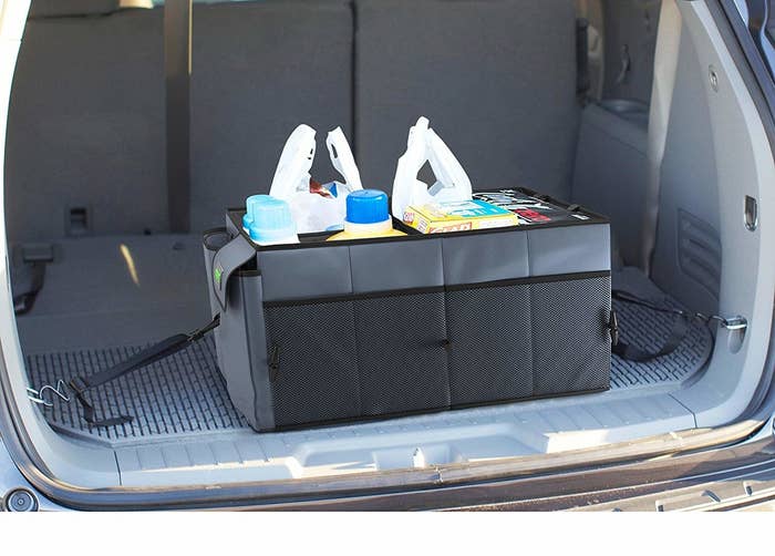 A large rectangular fabric box in the back of a car filled with groceries There are straps on each side that are connected to clips on the sides of the car