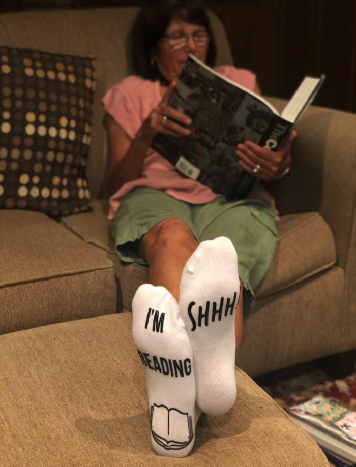 woman reading a book with her feet propped up and wearing the socks that say &quot;Shhh! I&#x27;m reading&quot; on the bottom of the feet