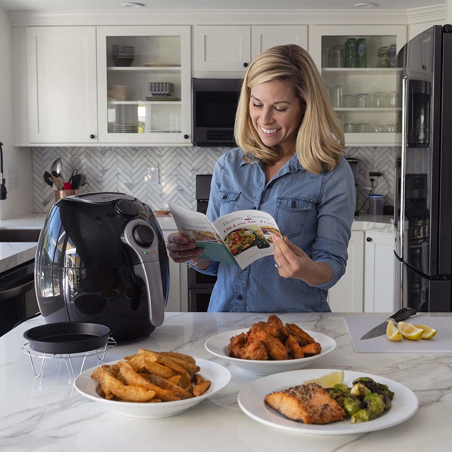 Model looking at the air fryer, reading the guide, with plates of air fried fries, wings, and salmon and brussels sprouts on the counter