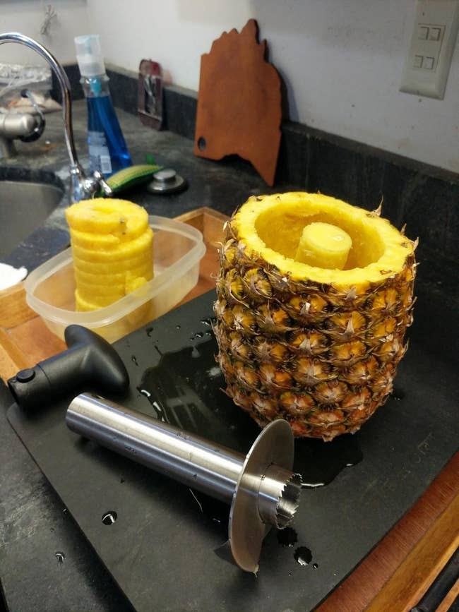 The tool on a counter with the cored pineapple shell and spiral-cut fruit in a container