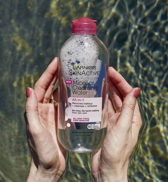 A pair of hands that are half submerged in water holding a bottle of the micellar water.