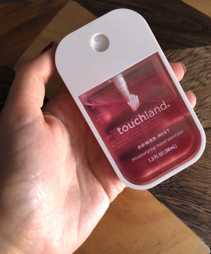 A hand holds the sanitizer container. The container has a rectangular shape (like a cell phone but smaller) and the front is a transparent pink so the liquid inside (and how much you have left) is visible. It also says the brand name &quot;Touchland.&quot;