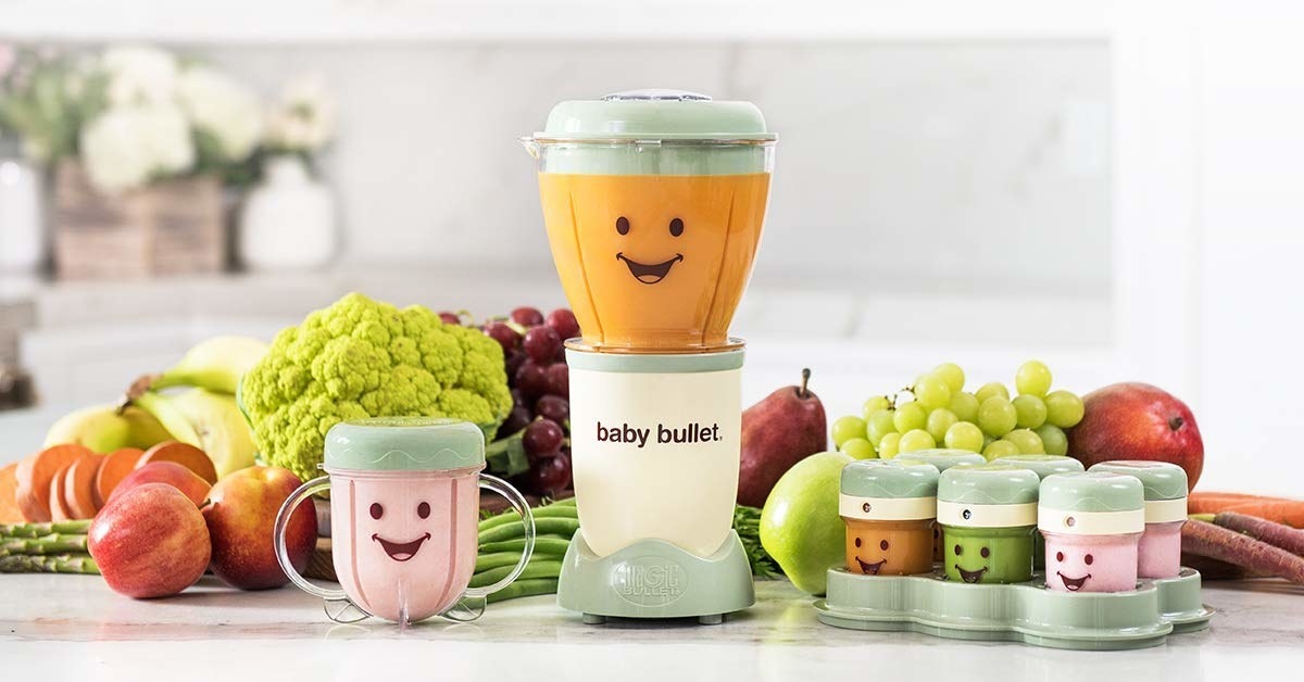 A small blender is sitting on a table with piles of fresh fruit and vegetables It has a cup with a lip next to it and a set of six small containers nearby