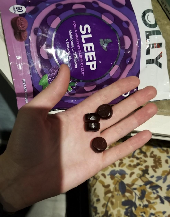 There is a hand holding four gummies. They are a deep dark purple. In the background is a bag that holds the Olly Sleep gummies.