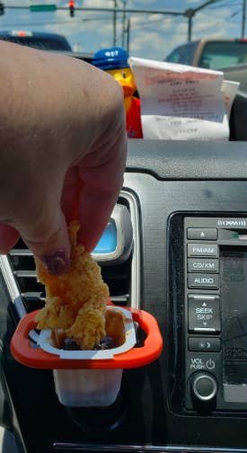 A reviewer dipping a chicken tender into a sauce cup stored in the red holder clipped to a car air vent