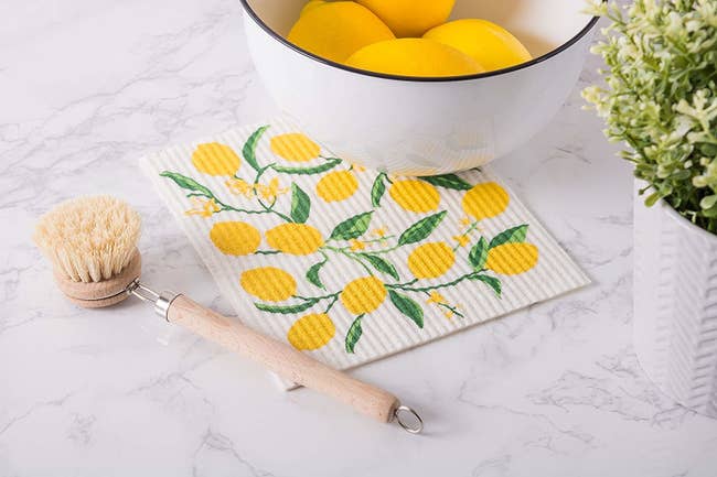 Lemon-patterned white dish cloth resting next to dish scrubber and bowl of lemons 