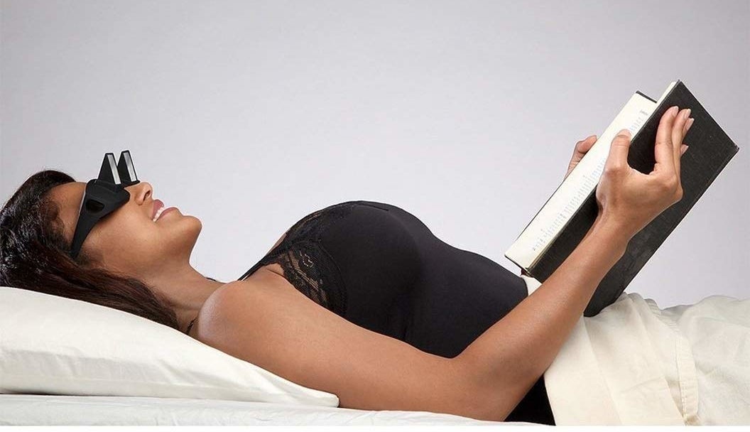 A model lays on her back in bed wearing black glasses that have a pyramidal attachment to the &quot;lens&quot; with the base facing a 90 degree angle downward to the book that she holds open over her stomach