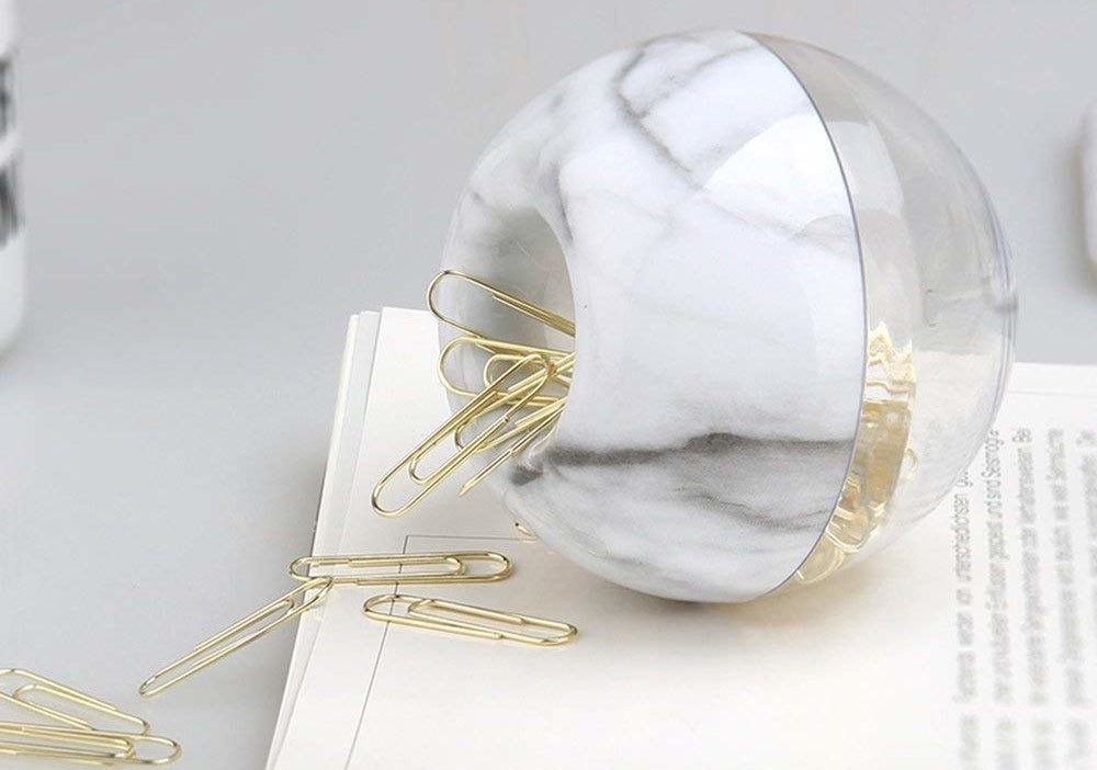 A small plastic ball with a hole in the top lying on its side on top of a book It&#x27;s filled with paper clips and some are falling out