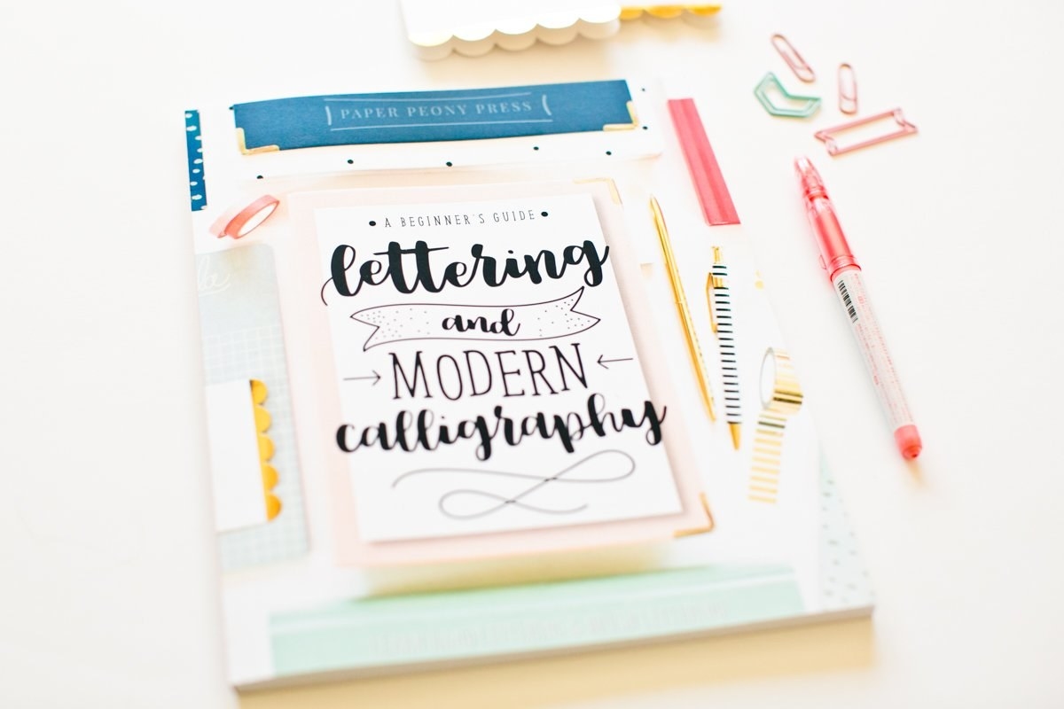 A small craft book called A Beginner&#x27;s Guide to Lettering and Modern Calligraphy on a desk with a pen and paper clips beside it