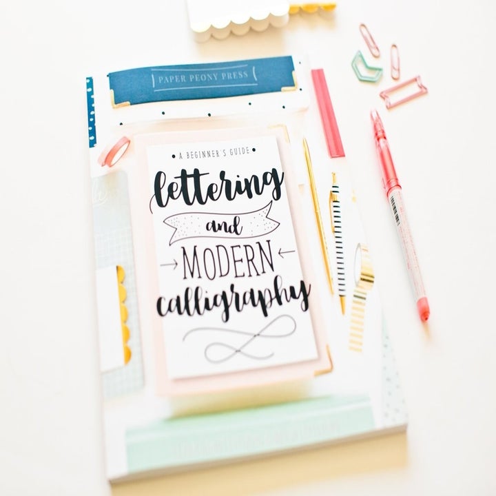 A small craft book called A Beginner's Guide to Lettering and Modern Calligraphy on a desk with a pen and paper clips beside it