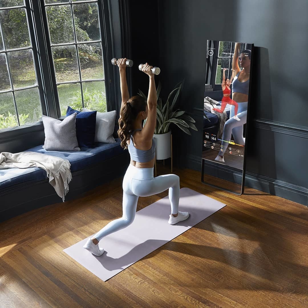 The rectangle-shaped mirror on a wall with a model on a yoga mat mirroring what the trainer is doing on the screen