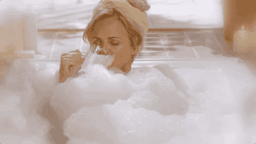 A gif of Amy Sedaris looking relaxed sipping some beer in a bubble bath