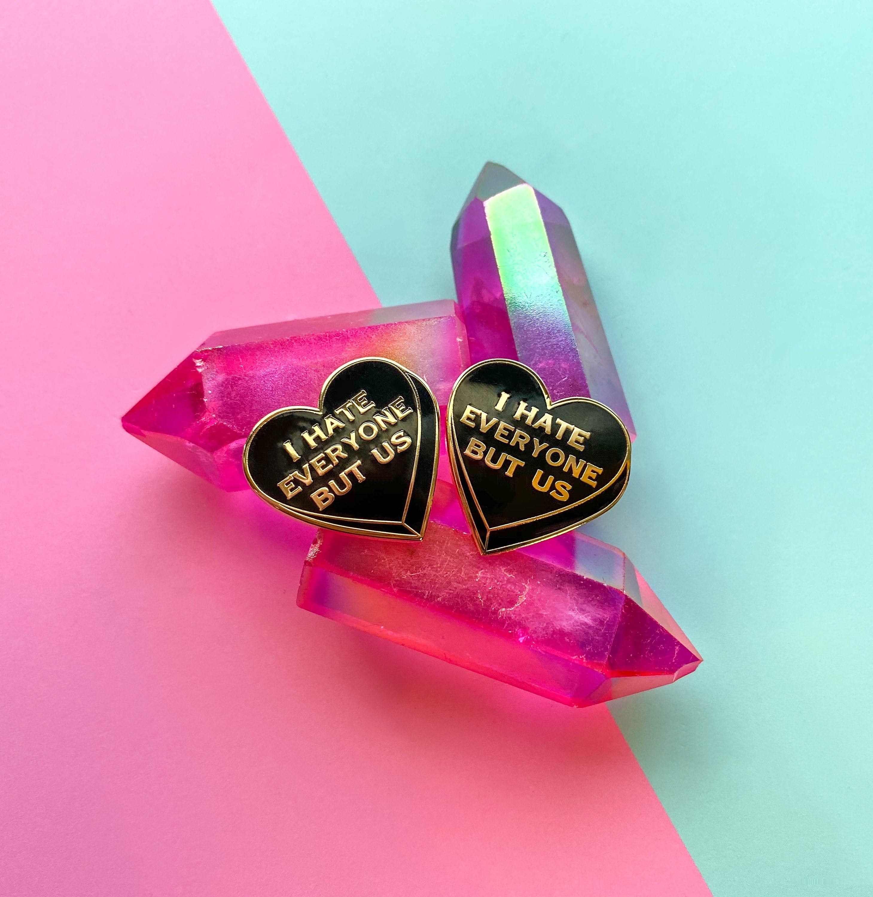 two black heart-shaped pins with &quot;i hate everyone but us&quot; written on them in gold