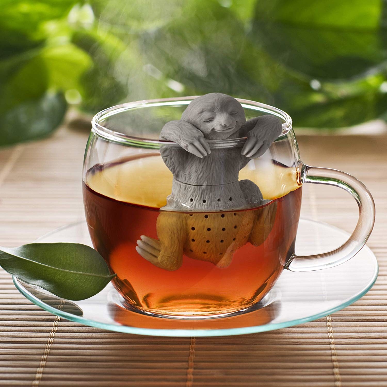 A clear glass saucer is holding a clear glass mug. Inside of the mug, you can see tea and there is a gray sloth tea infuser that is hanging from the rim (with its &quot;paws&quot;) and into the water for the tea.