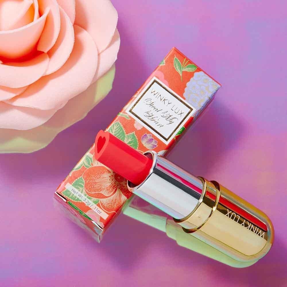 A gold tube of lipstick is open, showing a heart-shaped red lipstick, is propped on its floral box on a purple surface next to a pink rose. 