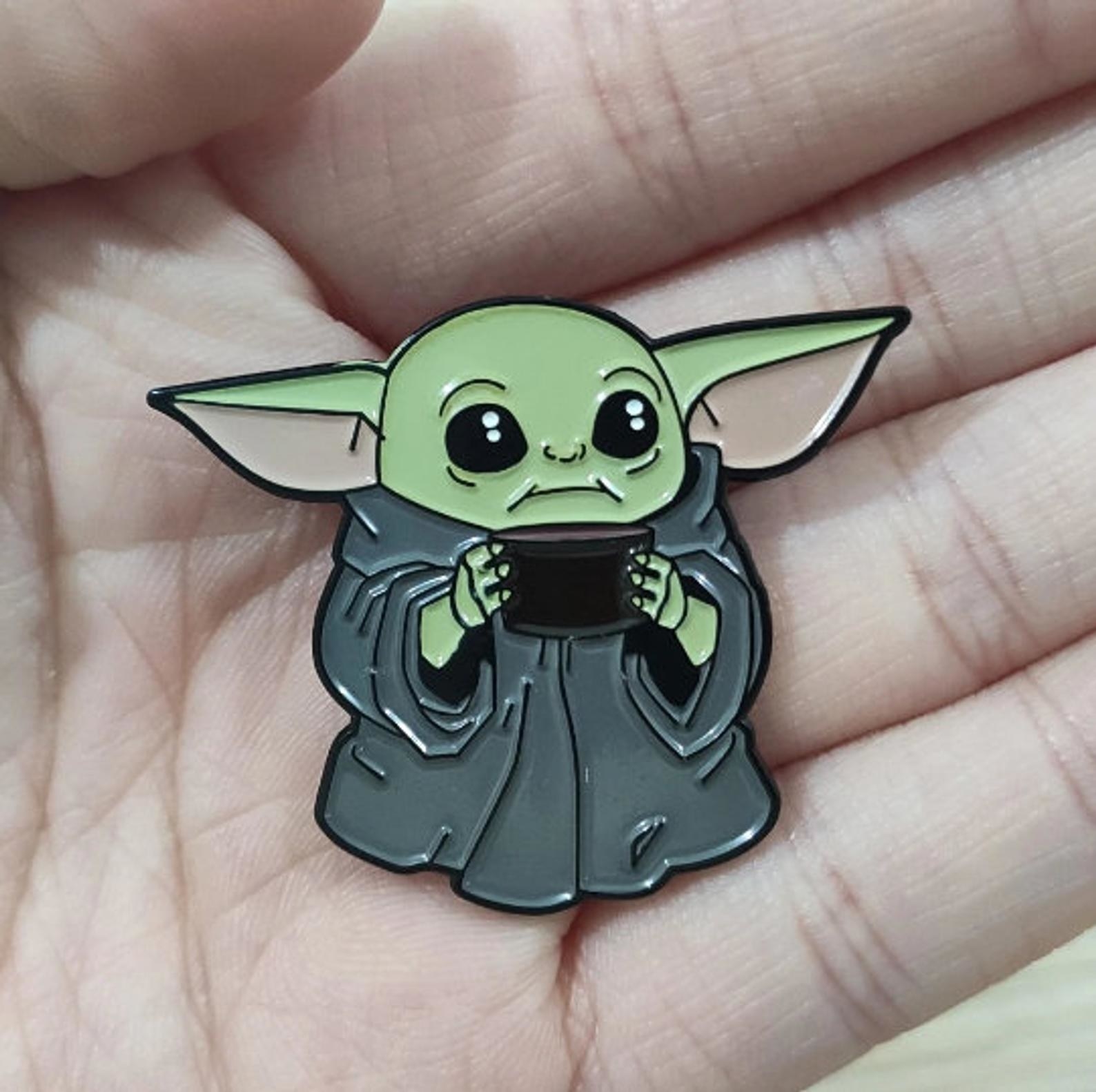 An enamel pin with an illustration of Baby Yoda iconically holding a cup of tea. 