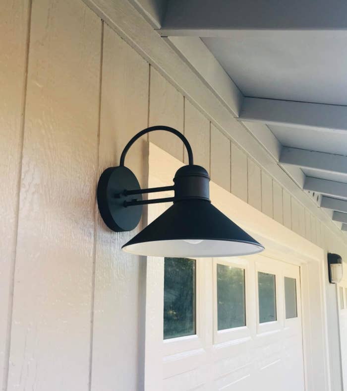 Reviewers pic of the black wall sconce with circle base and a curved top bar attached to the outside of a house.