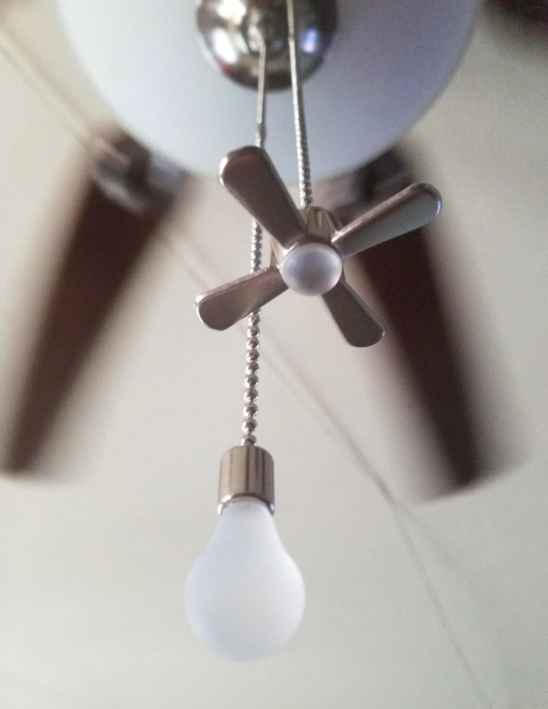 A string with a lightbulb on one side and little fan on the other side hanging from a ceiling fan.