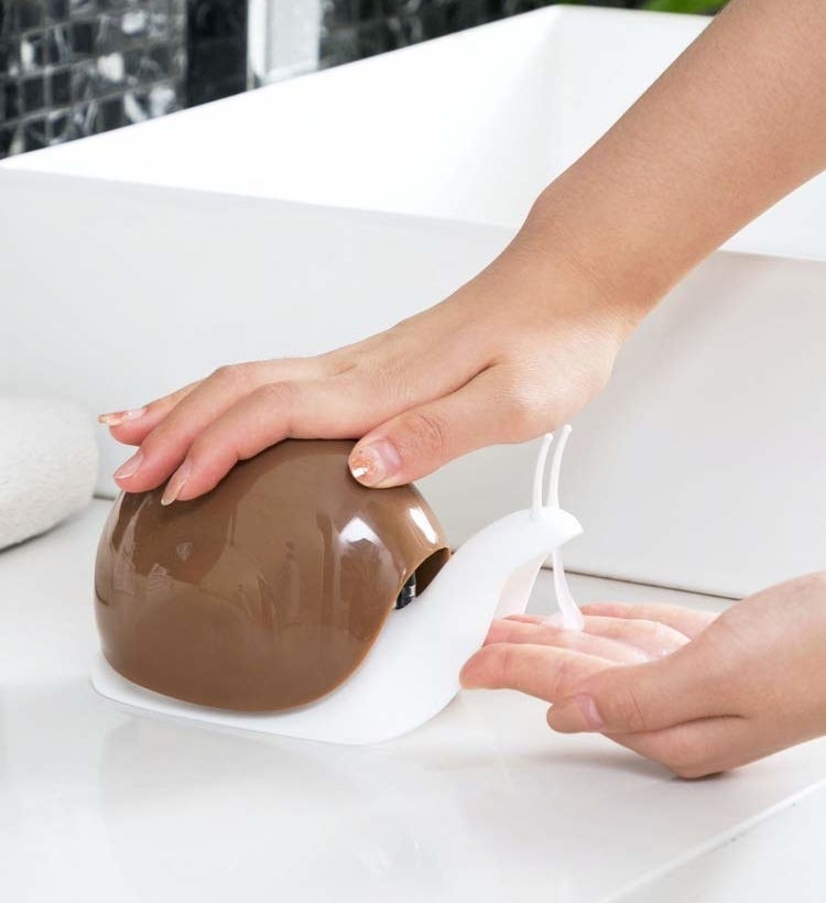 A hand pressing down on the shell of the brown and white snail dispenser, which is dispensing soap from the snail&#x27;s mouth