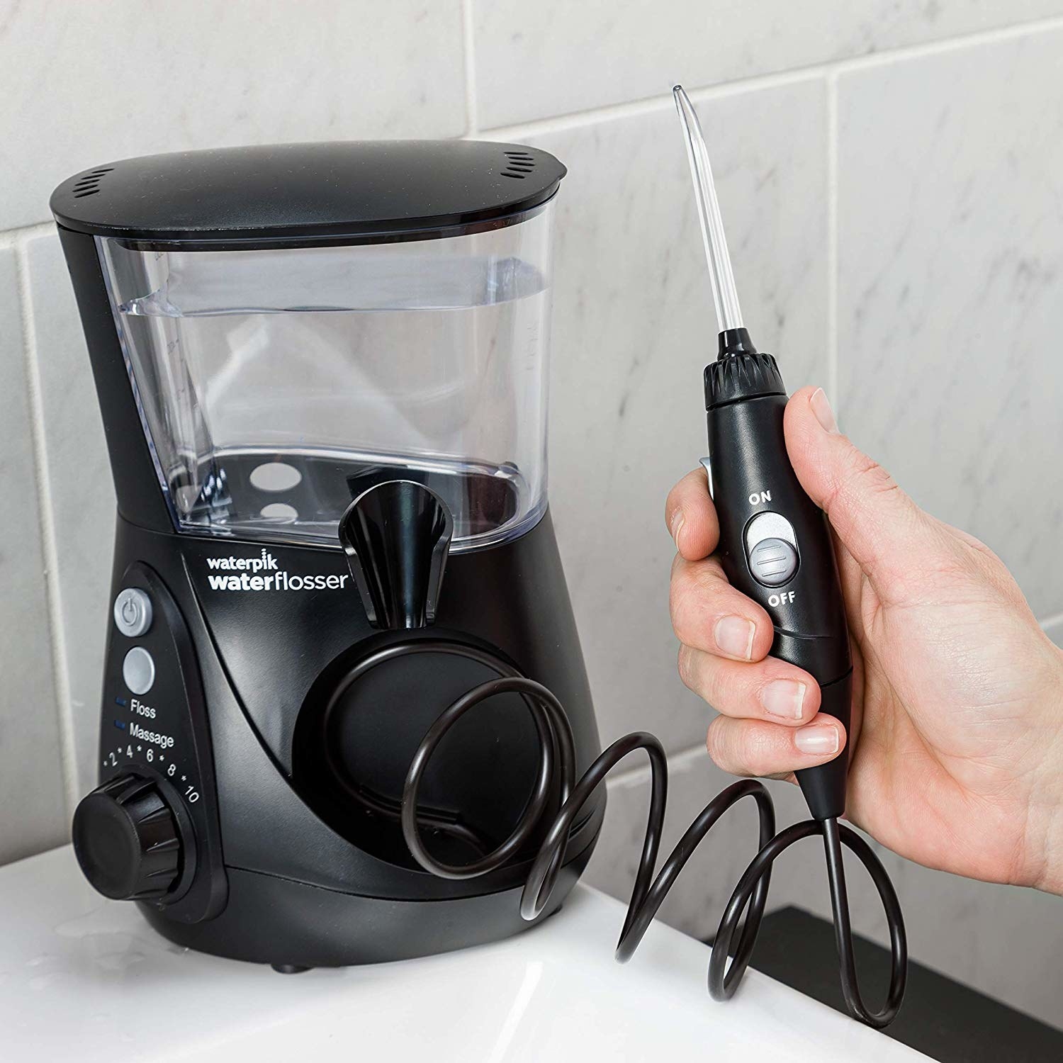 Water flosser in hand. The flosser has a handle with a narrow tip and a wire connecting it to a water reservoir 