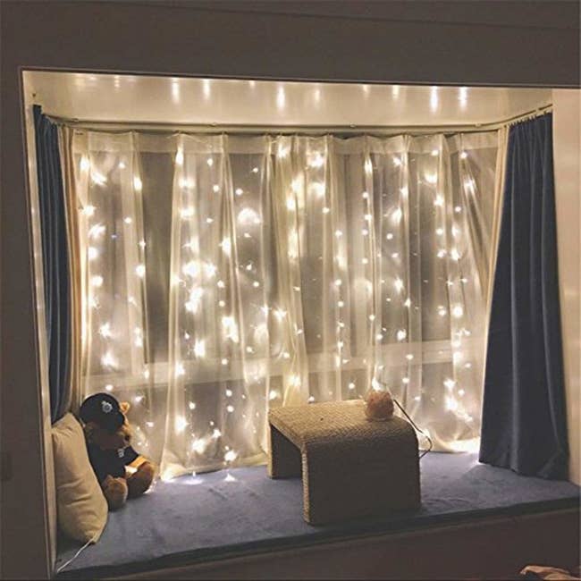 A dark window with sheer curtains and yellow fairy lights strung up illuminating it 