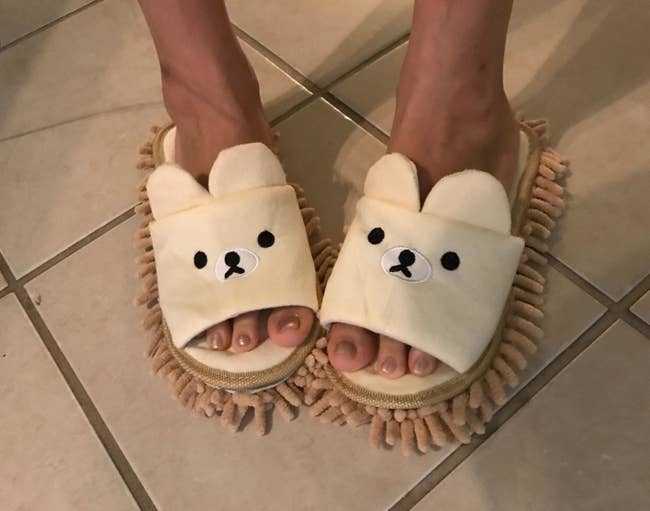 Reviewer wearing the slippers in beige with a face and ears on the top and light brown mop on the bottom.
