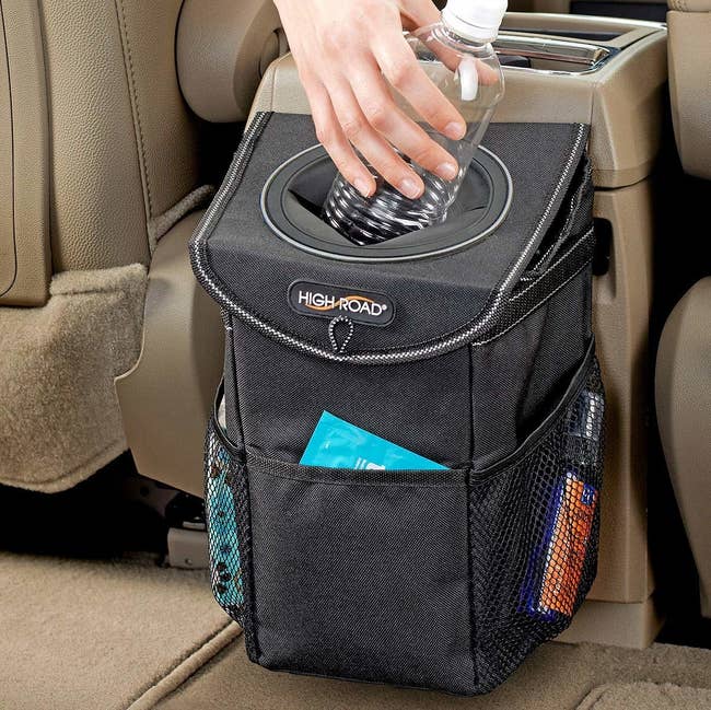 A small black trash can hooked to the back of a car console with pockets and a little hole for depositing trash 