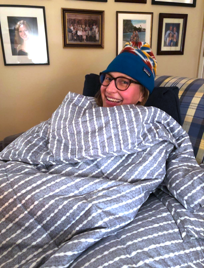 a reviewer snuggled up and smiling underneath a blue and white striped weighted blanket