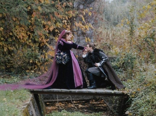 bride and groom wearing purple renaissance clothing on a small bridge in the woods. the groom kneels to kiss the bride&#x27;s hand.