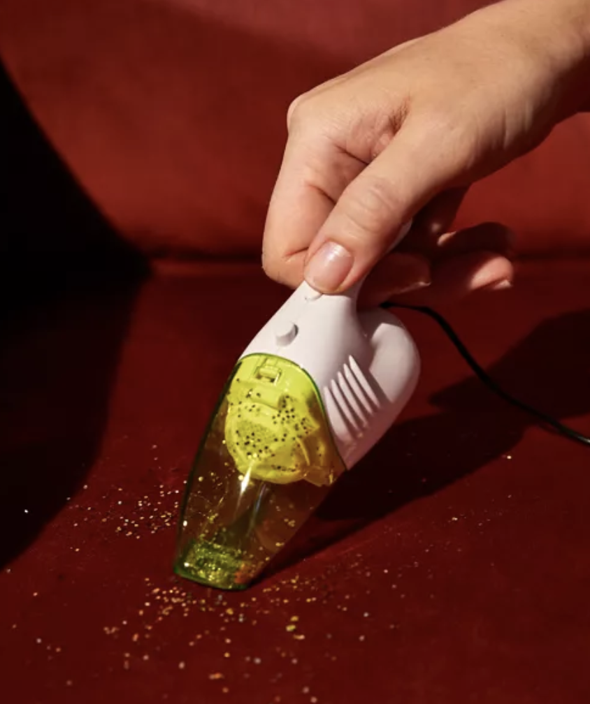 hand holds tiny yellow handheld vacuum sucking up glitter from a couch