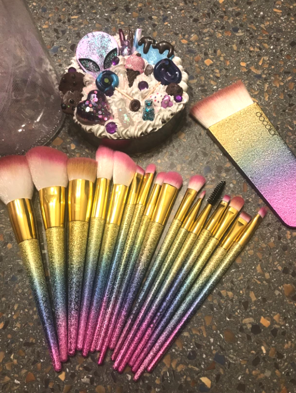 Glittery makeup brushes with an ombre design on their handles featuring pink, blue, and gold. They are laid out on a surface.