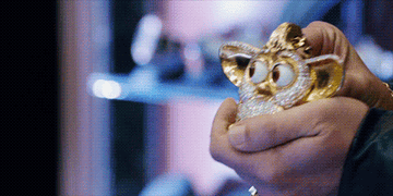 gif of adam sandler in uncut gems showing off a bejeweled furbee with moving eyes on a chain to a laughing crowd in his store