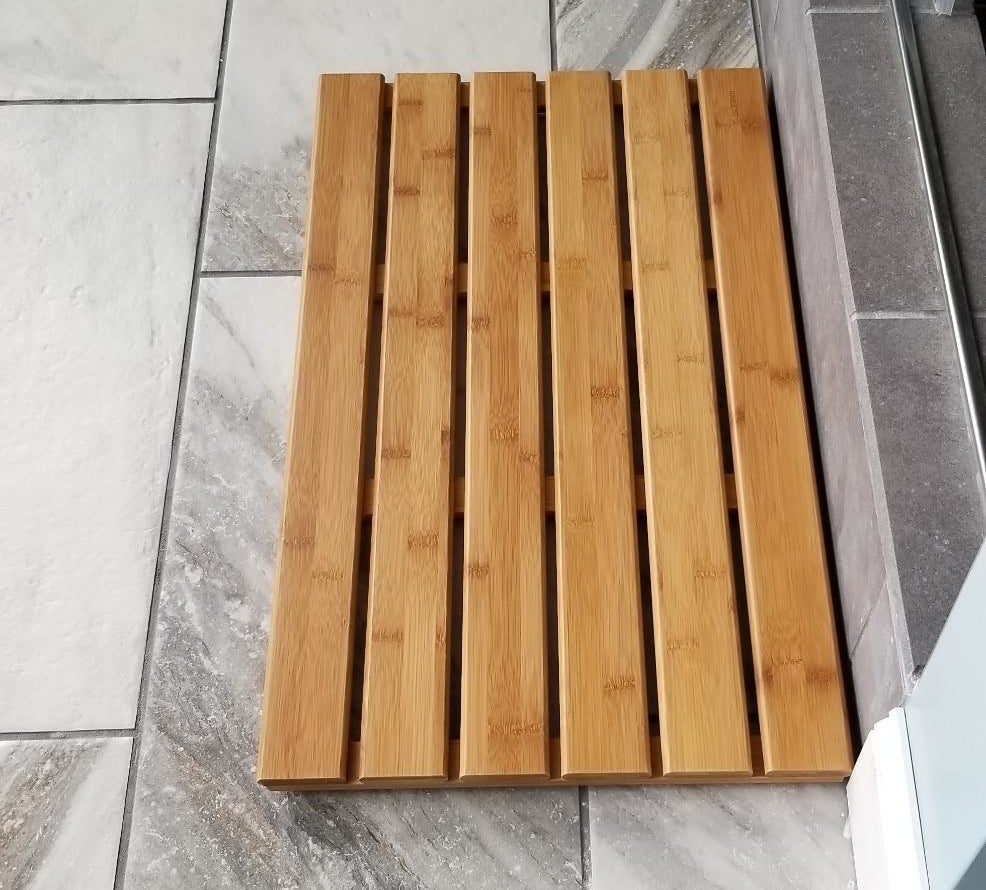 a reviewer photo of the slatted natural bamboo mat on the floor of their bathroom