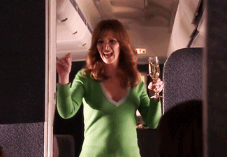 GIF of Lisa Kudrow walking down airplane aisle with glass of champagne
