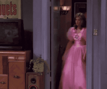 Jennifer Aniston walking in wearing a pufffy pink princess dress in a scene of the Friends sitcom series. She is clearly not feeling the dress.