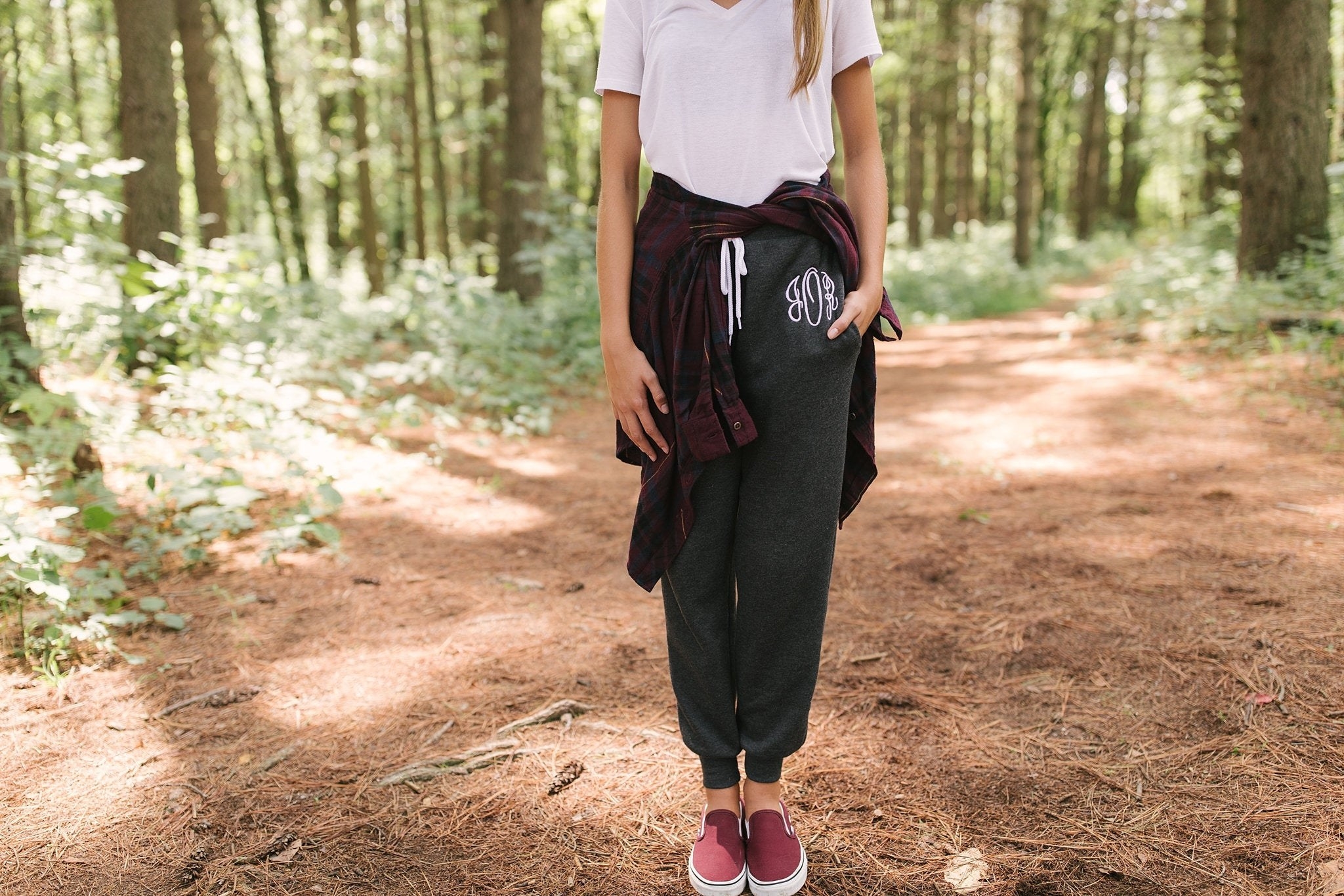 A model standing in the woods wearing the sweatpants with a T-shirt, a button-up shirt wrapped around the waist, and slip-on sneakers