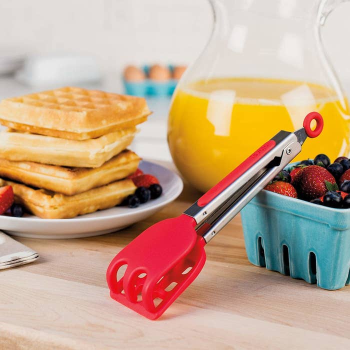 A pair of waffle tongs leaning against a carton of berries in front of a stack of waffles