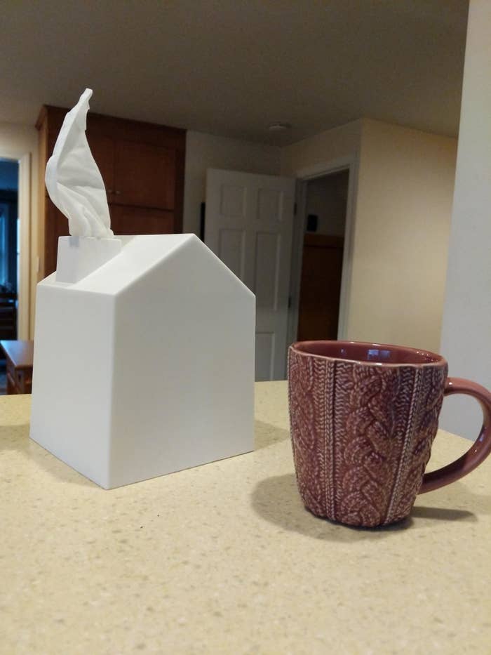 A white tissue box shaped like a house with a chimney that a tissue is coming out of, propped on a table 