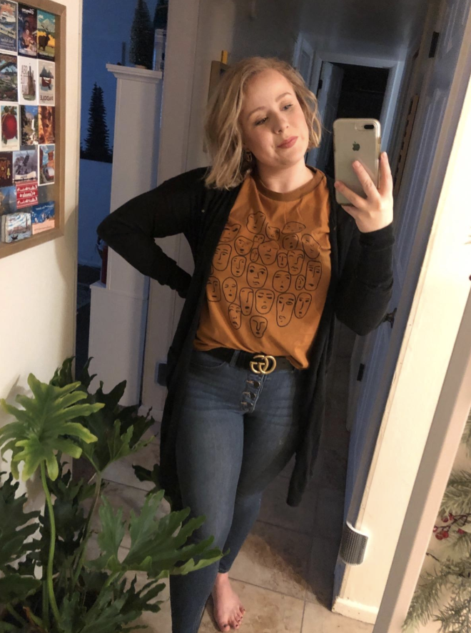 Reviewer wears button up skinny jeans, a black cardigan, and mustard yellow tee with artsy face illustrations
