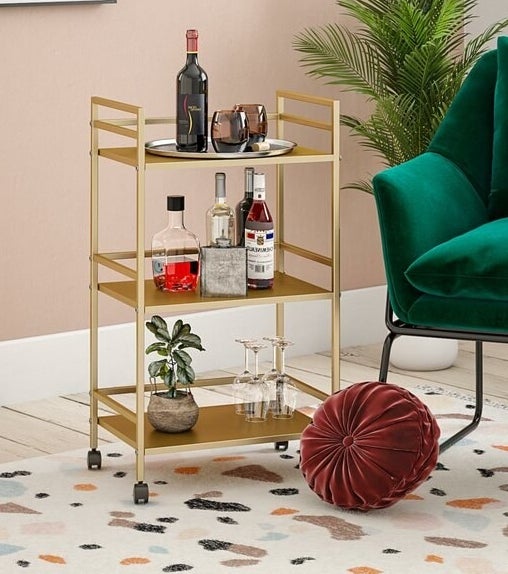 A gold metallic three-shelf bar cart on four caster wheels filled with bottles and glasses situated atop a colorful rug