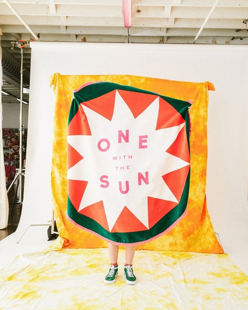 a person holding up a bright yellow towel with a green circle around an orange circle around a white star with the words &quot;one with the sun&quot; in the middle in pink